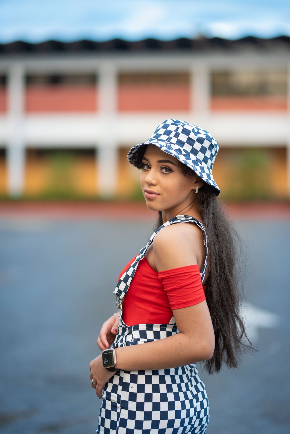 Young woman posing outdoors in stylish blue checkered bucket hat and matching overalls with red off-shoulder top.