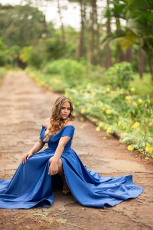 Young woman in elegant royal blue off-shoulder gown sitting gracefully on a rustic dirt path surrounded by lush greenery.