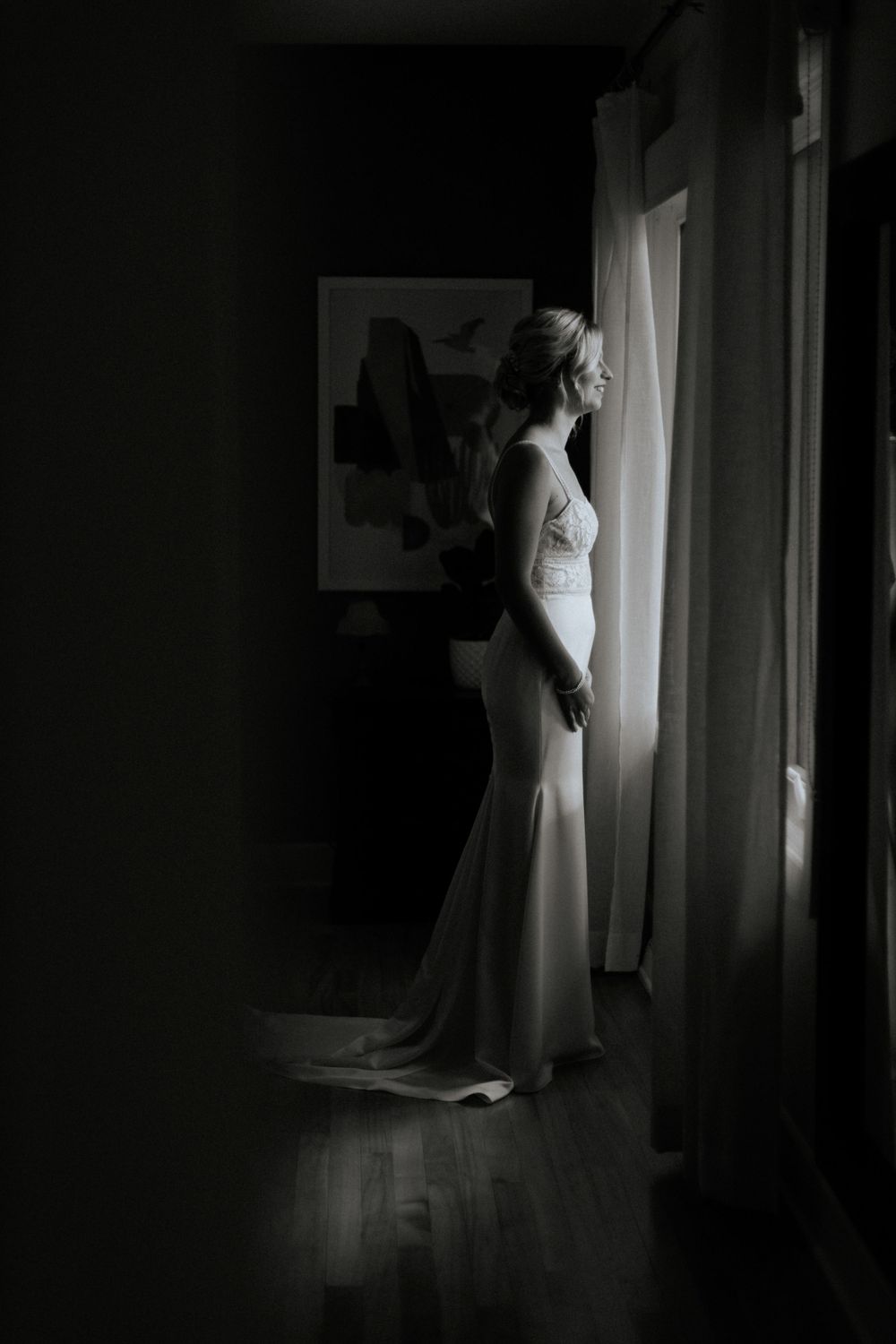 Silhouette of a bride in a fitted wedding gown standing by a window in dramatic lighting.