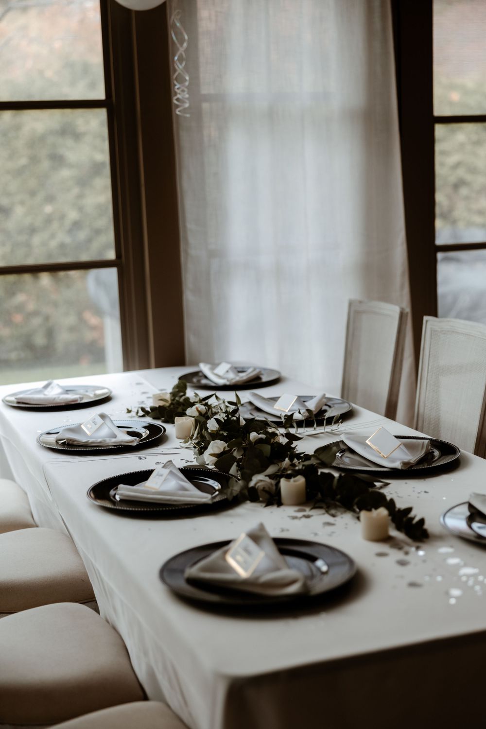 Elegant table setting with black chargers and white linens in front of sheer curtained windows.