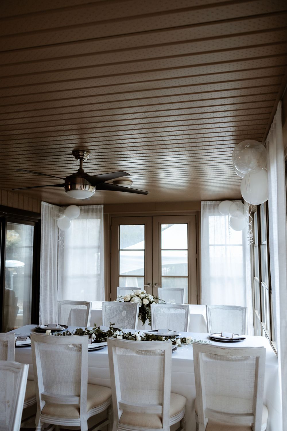 Bright sunlit dining room with white chairs and table settings under wooden ceiling with fan.