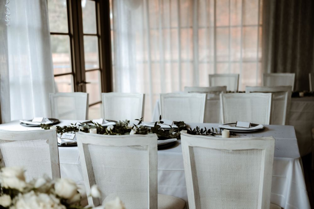 White dining chairs and table settings with greenery centerpieces in an elegant reception space.