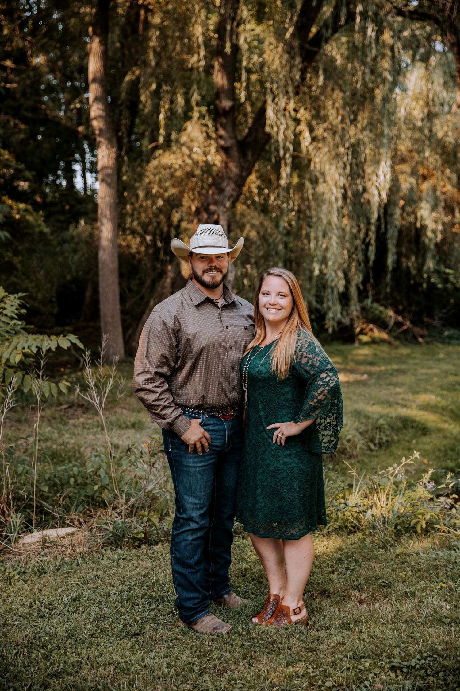 A couple poses outdoors under willow trees, with the man wearing a cowboy hat and the woman in a green dress.