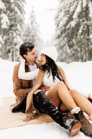 Couple sharing an intimate moment while sitting on a blanket in a snowy forest setting.