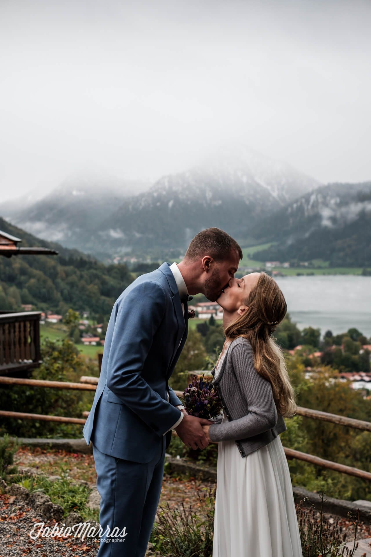 Brautpaar küsst sich innig vor der nebligen Bergkulisse der Alpen am Schliersee in Bayern.