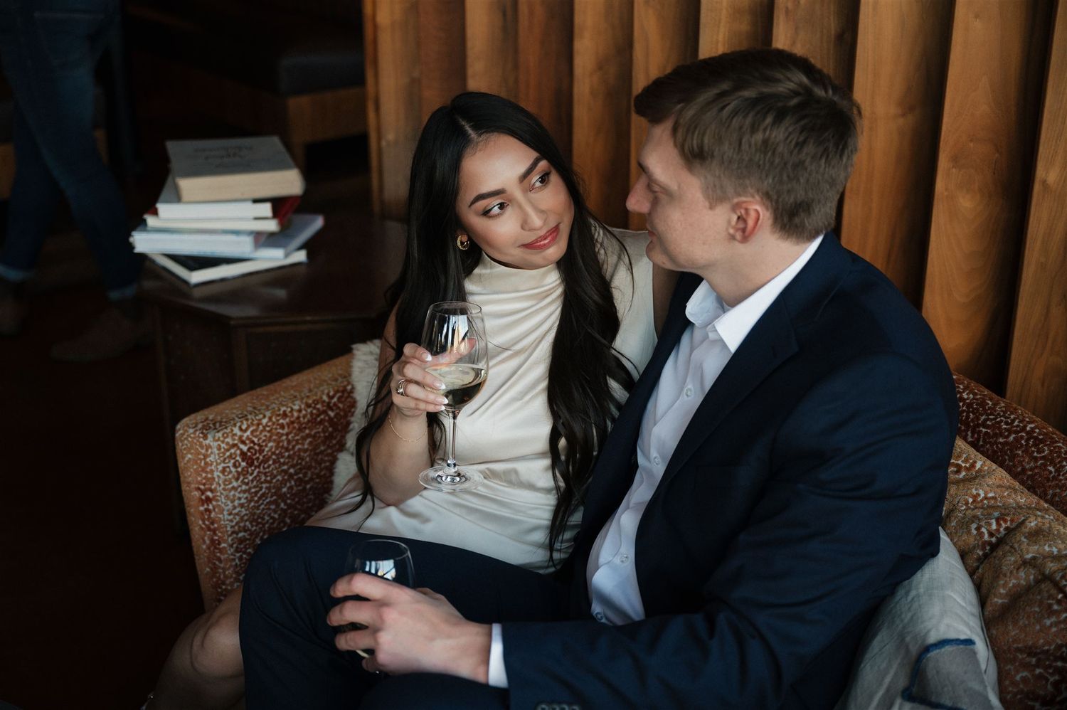 Intimate couple enjoying wine together on vintage leather sofa against wooden wall backdrop.