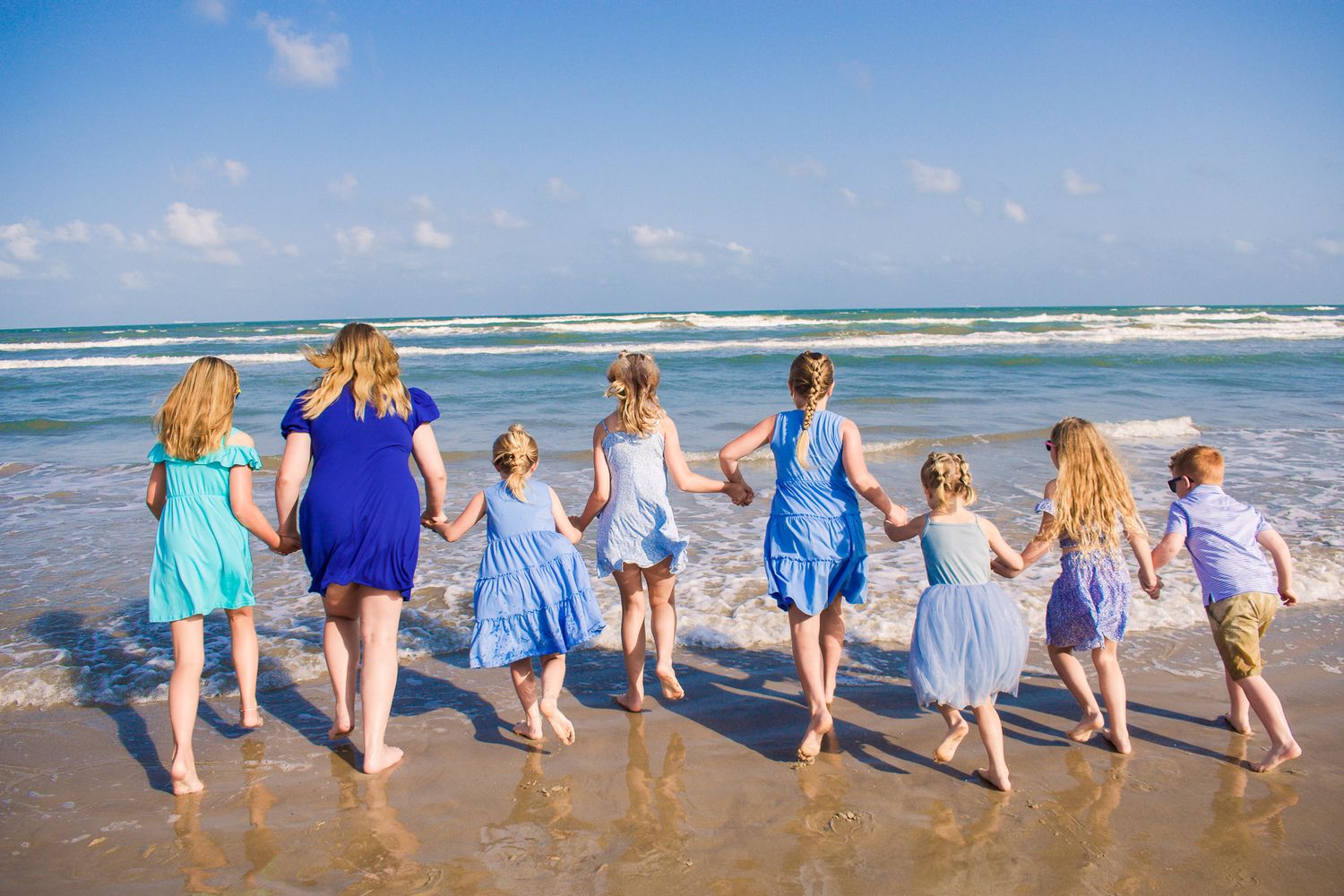 Group holding hands while running together on beach in coordinated blue dresses.