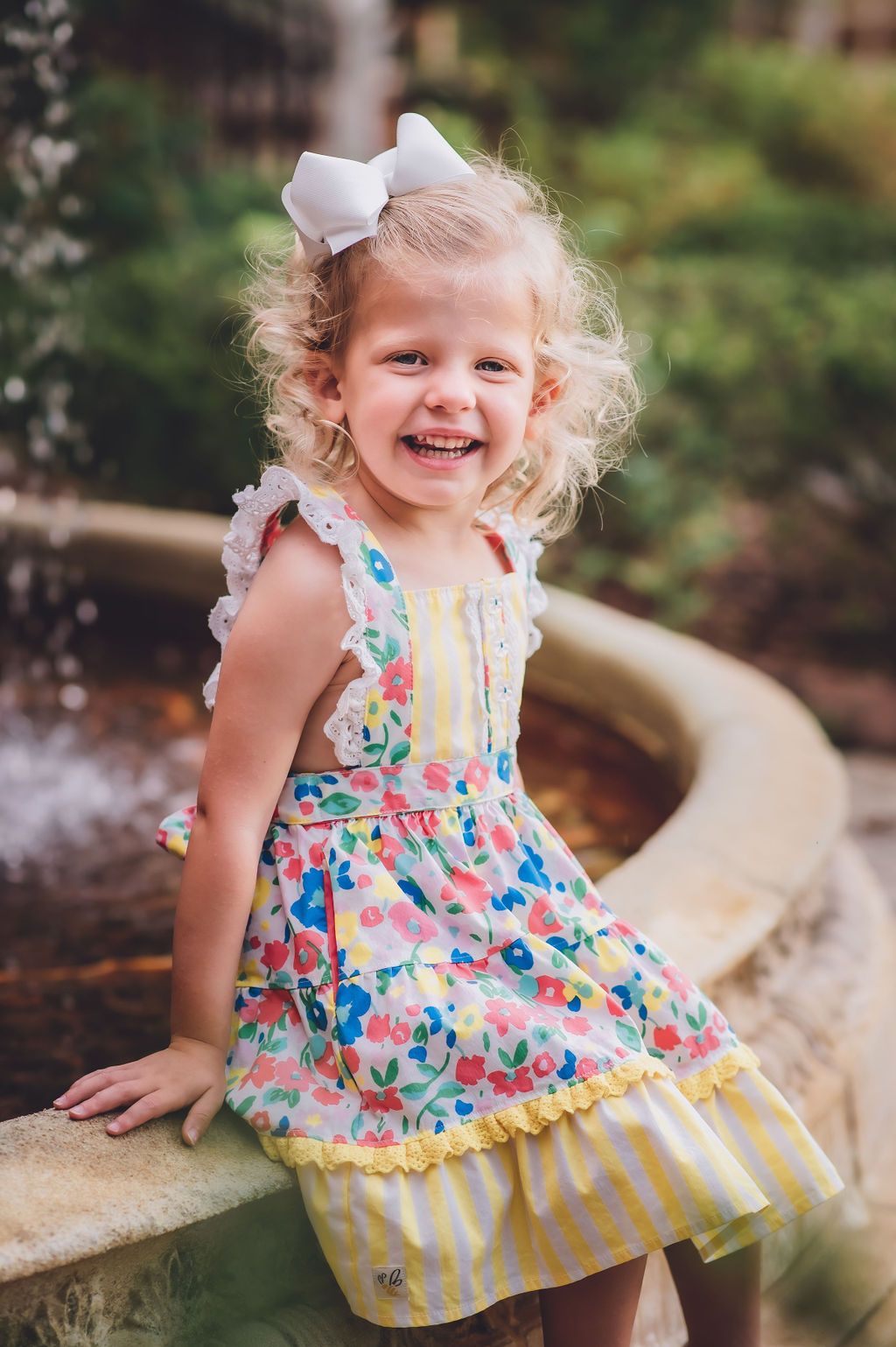 3-year-old sits on side of fountain in floral Wild Flowers brand apron dress for her birthday photo session