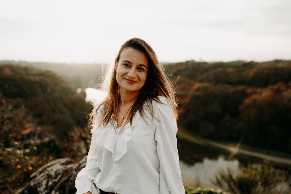 Woman in white blouse smiling with autumn scenery and river view in background.