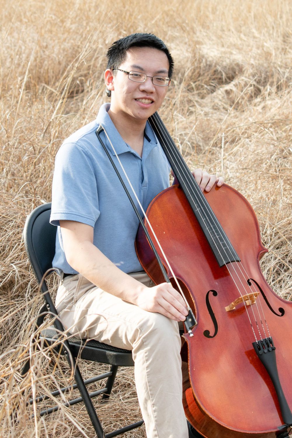 A musician poses with a cello while seated on a folding chair in tall grass.