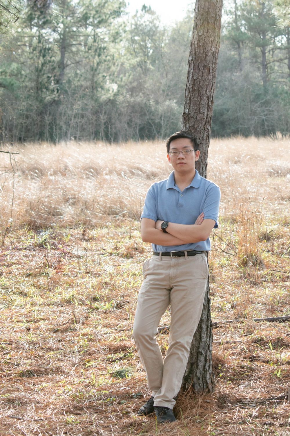 A musician in a blue polo stands casually against a pine tree in a natural setting.