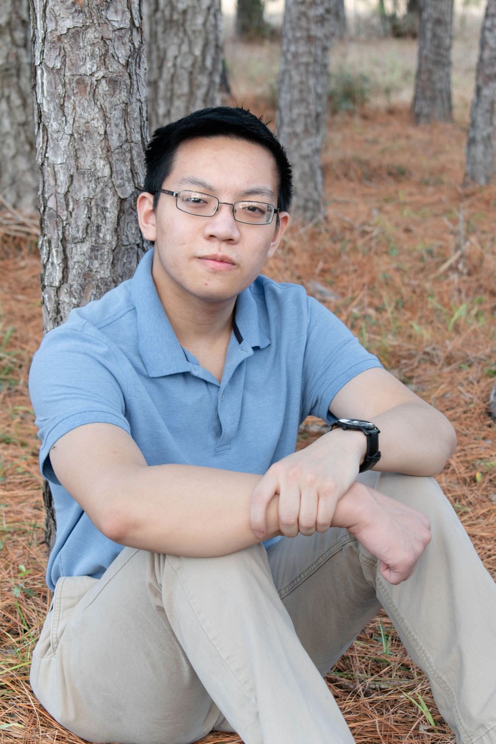 A musician in casual attire sits cross-legged against a tree in a wooded setting.