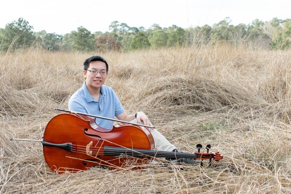 A cellist sits with their instrument in a field of dried grass during golden hour.
