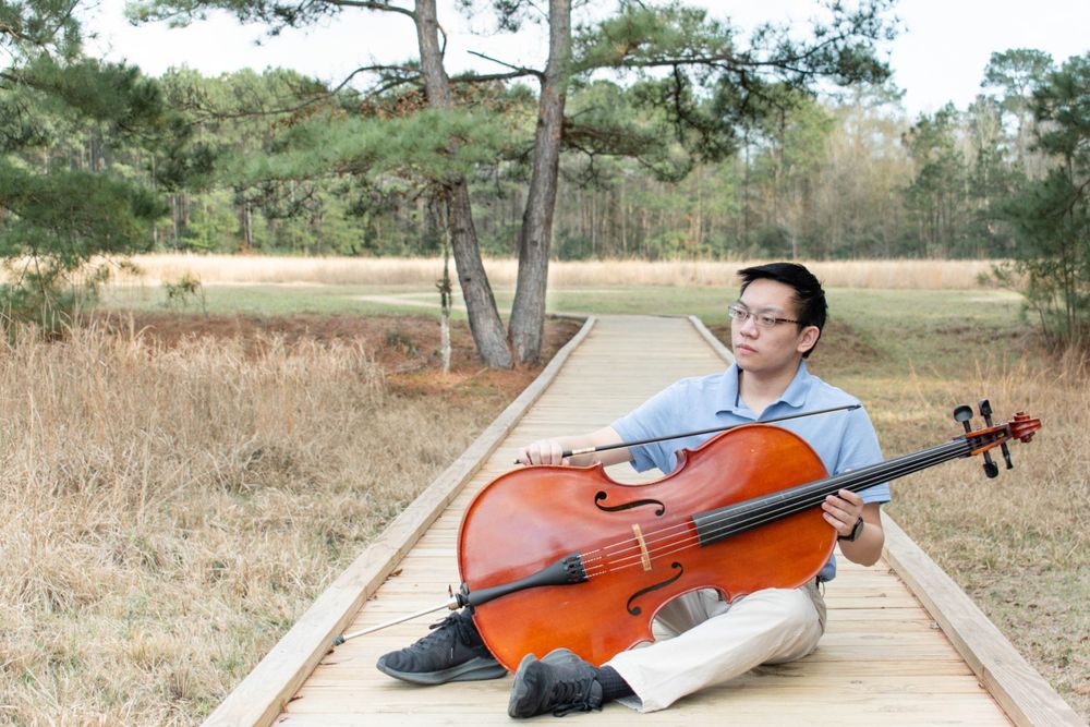 A cellist poses with their instrument on a wooden walkway surrounded by tall grass.