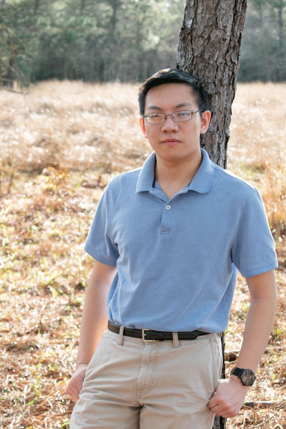 A musician in a blue polo and khakis stands against a tree trunk in an autumn field.