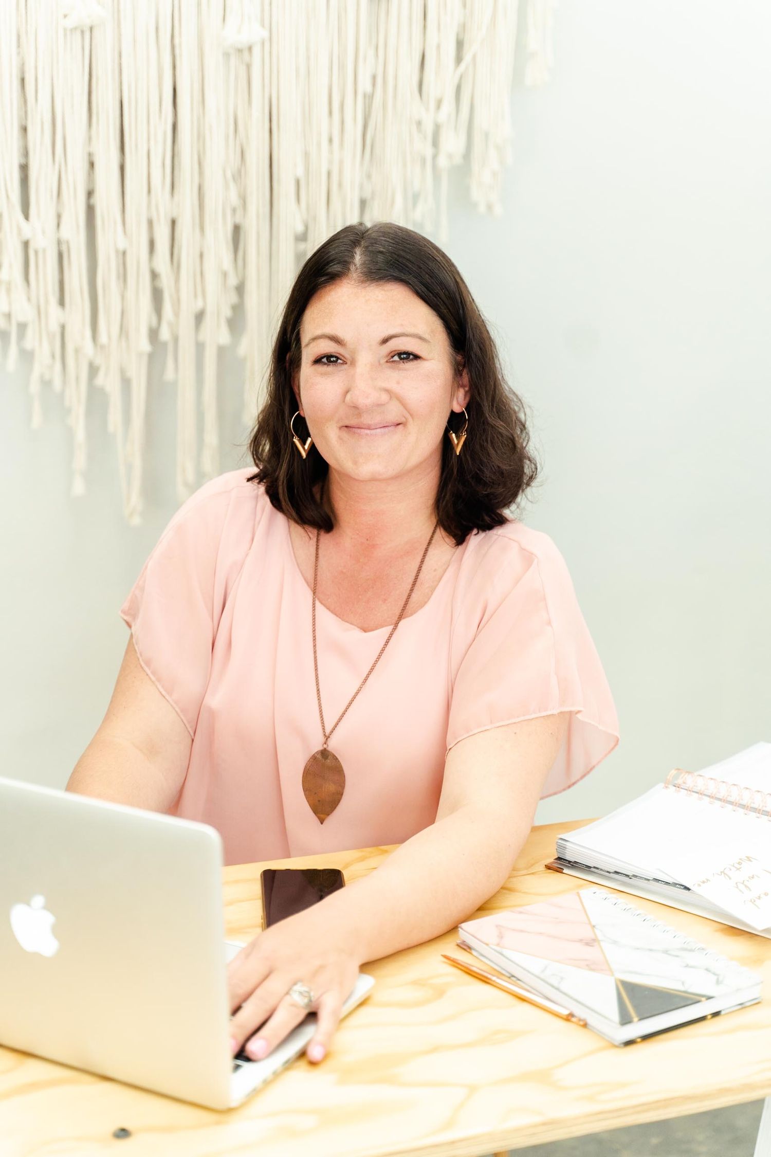 Person in pink blouse with pendant necklace working on MacBook at wooden desk.