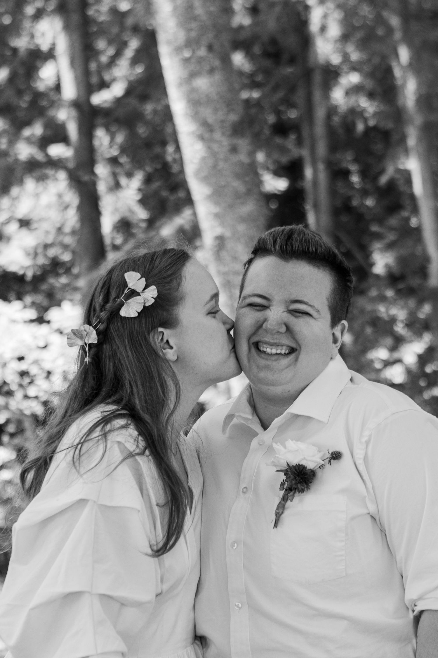 A queer married couple kisses one another on the cheek on their wedding day, photographed in black and white