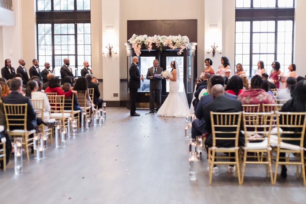 Elegant indoor wedding ceremony with guests seated in gold chairs and floral arch at the altar.