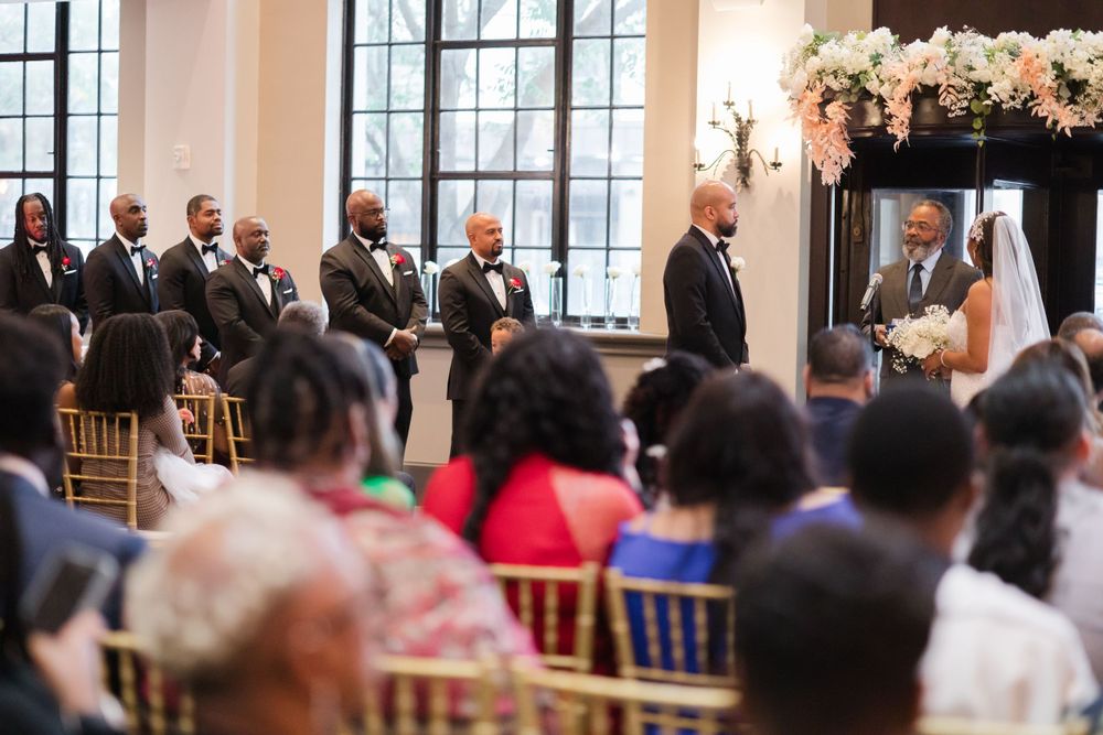 Wedding ceremony in a bright venue with tall windows and guests seated in gold chairs.