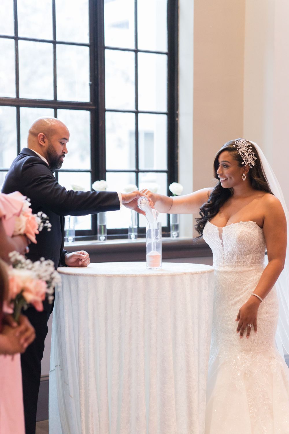 Wedding ceremony participants perform unity sand ritual at white-draped table near large windows.