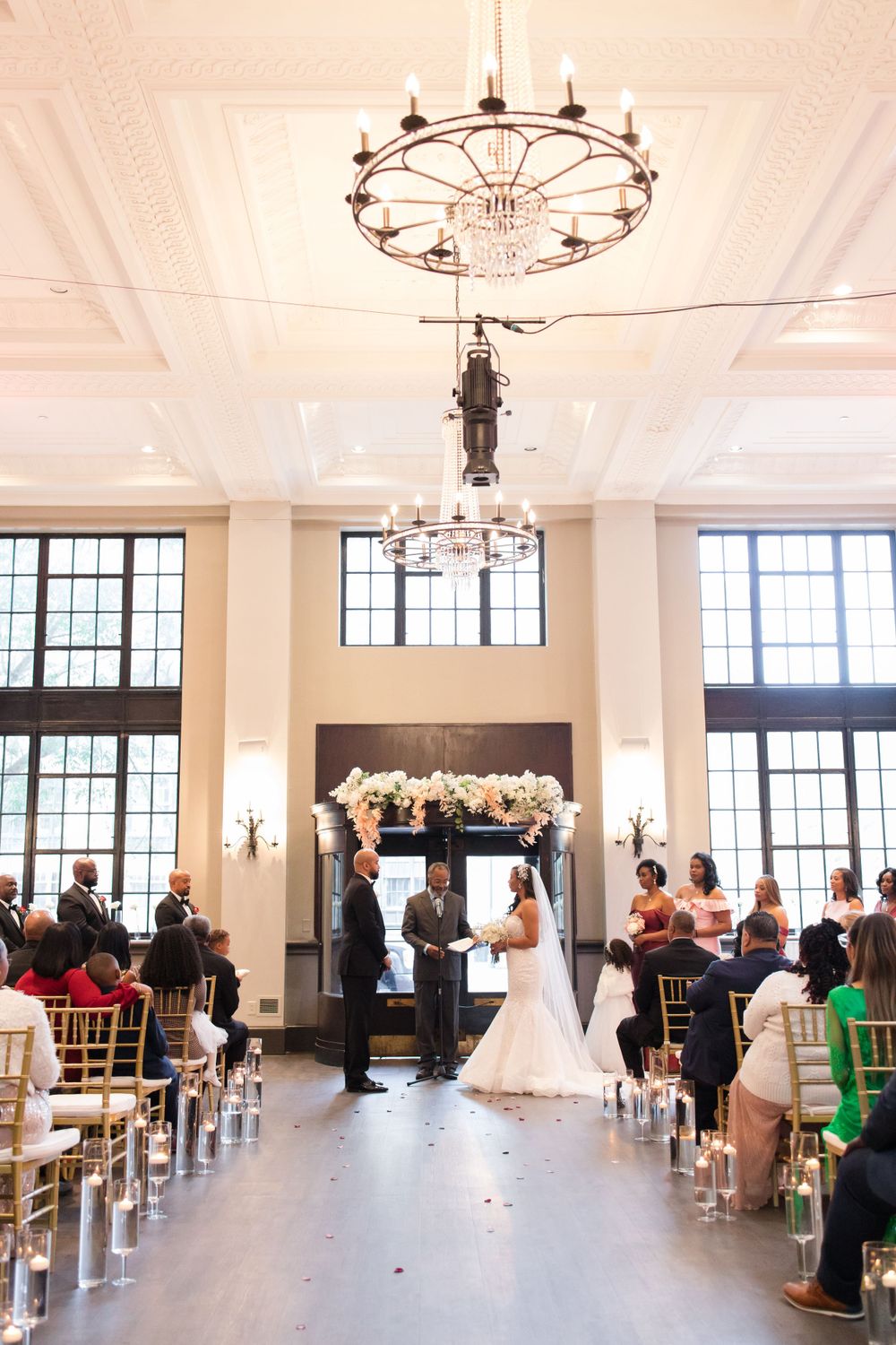 Wedding ceremony in elegant hall with grand chandelier and floral arch backdrop.