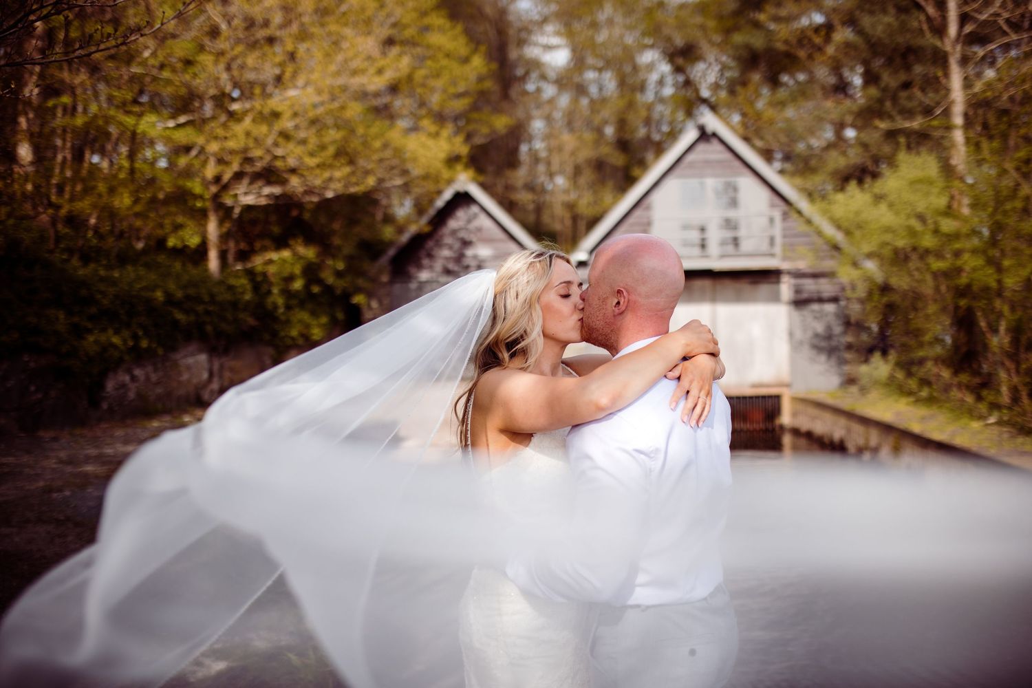Romantic wedding couple embracing with flowing veil against rustic building backdrop.