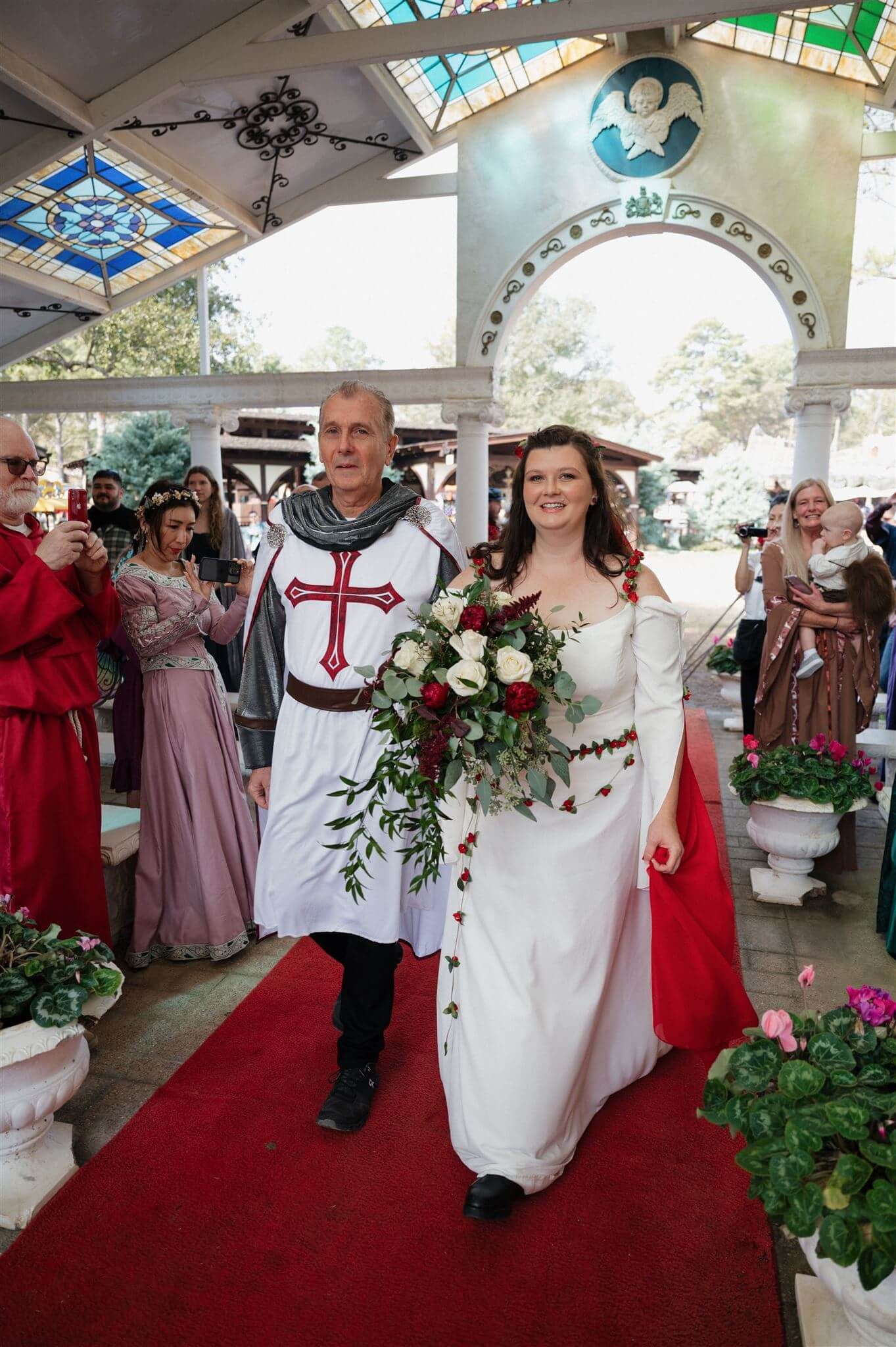 Medieval-themed wedding procession walks down red carpet under ornate gazebo with stained glass skylights.