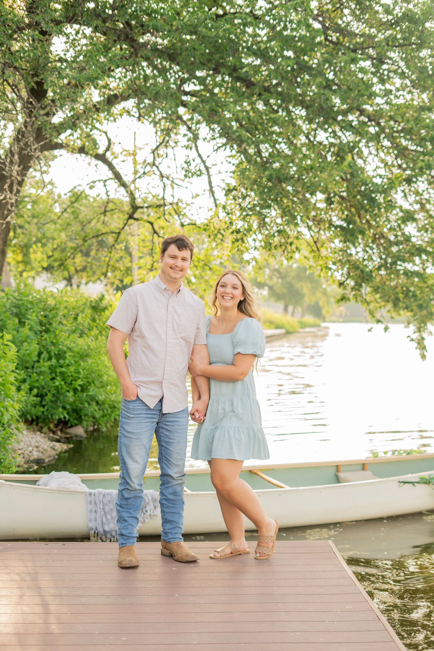 Couple smiling and holding hands on a dock beside a white canoe on a serene river.