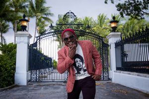 A person in a red blazer poses playfully in front of an ornate white gate with palm trees.