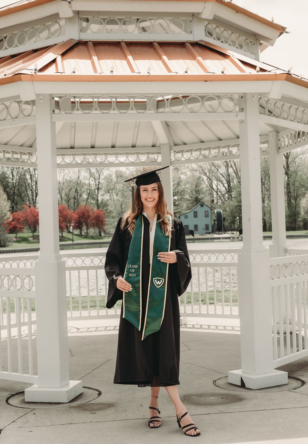 A graduate in cap and gown poses under a white gazebo with green graduation stole.