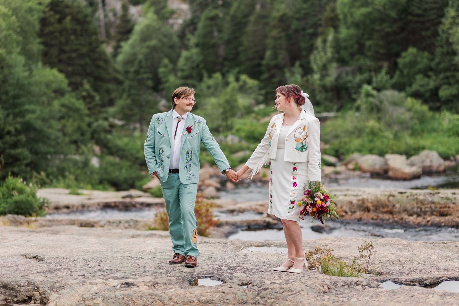 Casual wedding couple walks hand in hand on rocky mountain terrain.