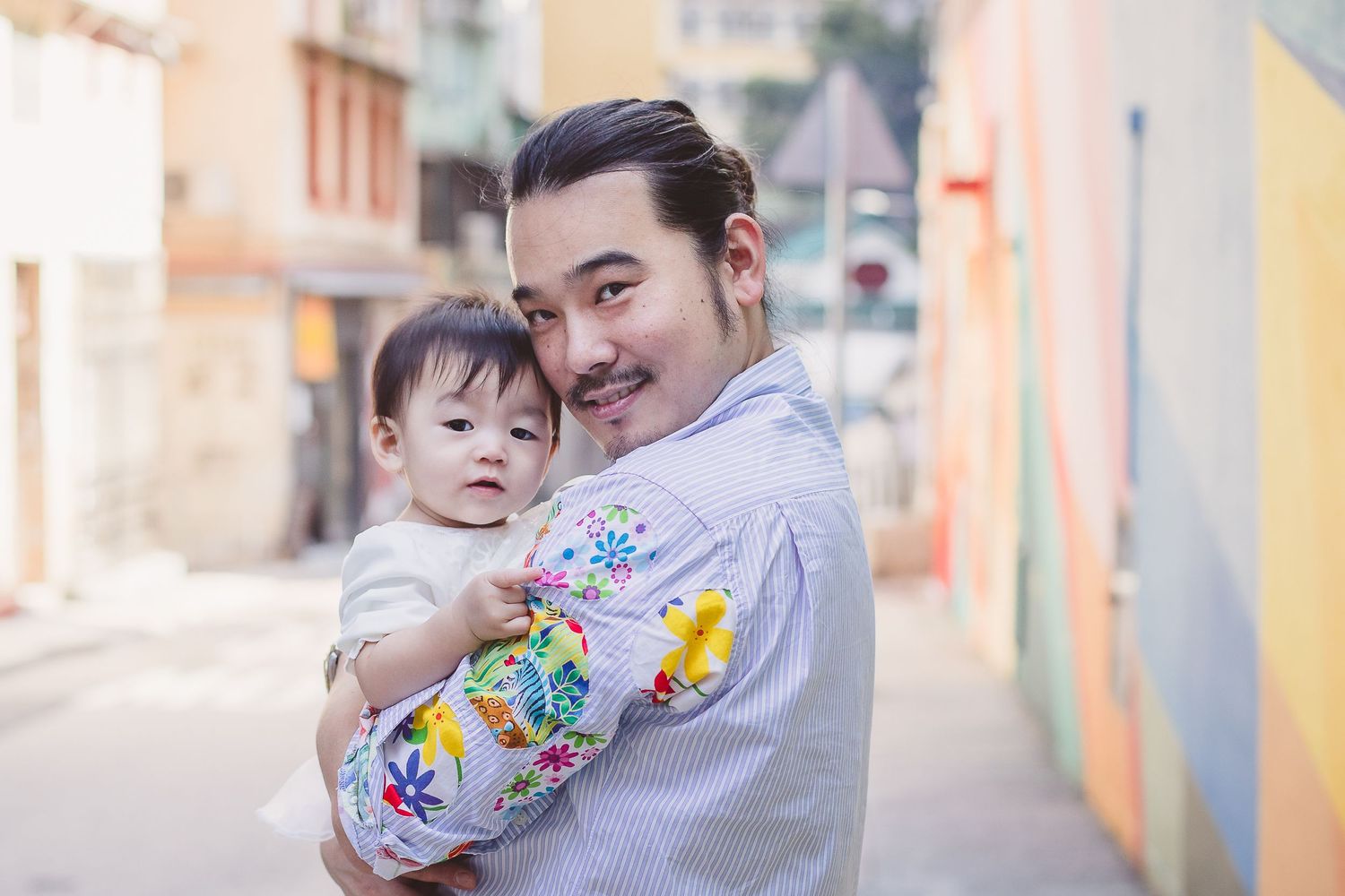 Parent holding smiling toddler in floral outfit against pastel colored urban backdrop.