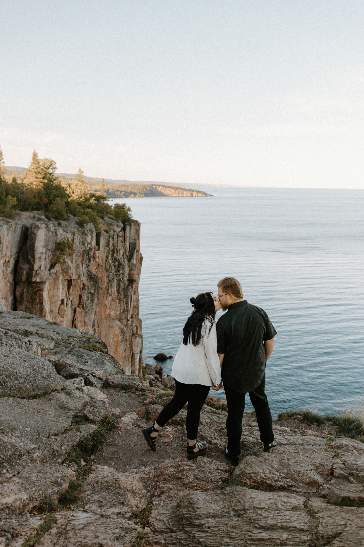 Two people stand on rocky cliffs overlooking a serene lake at sunset.