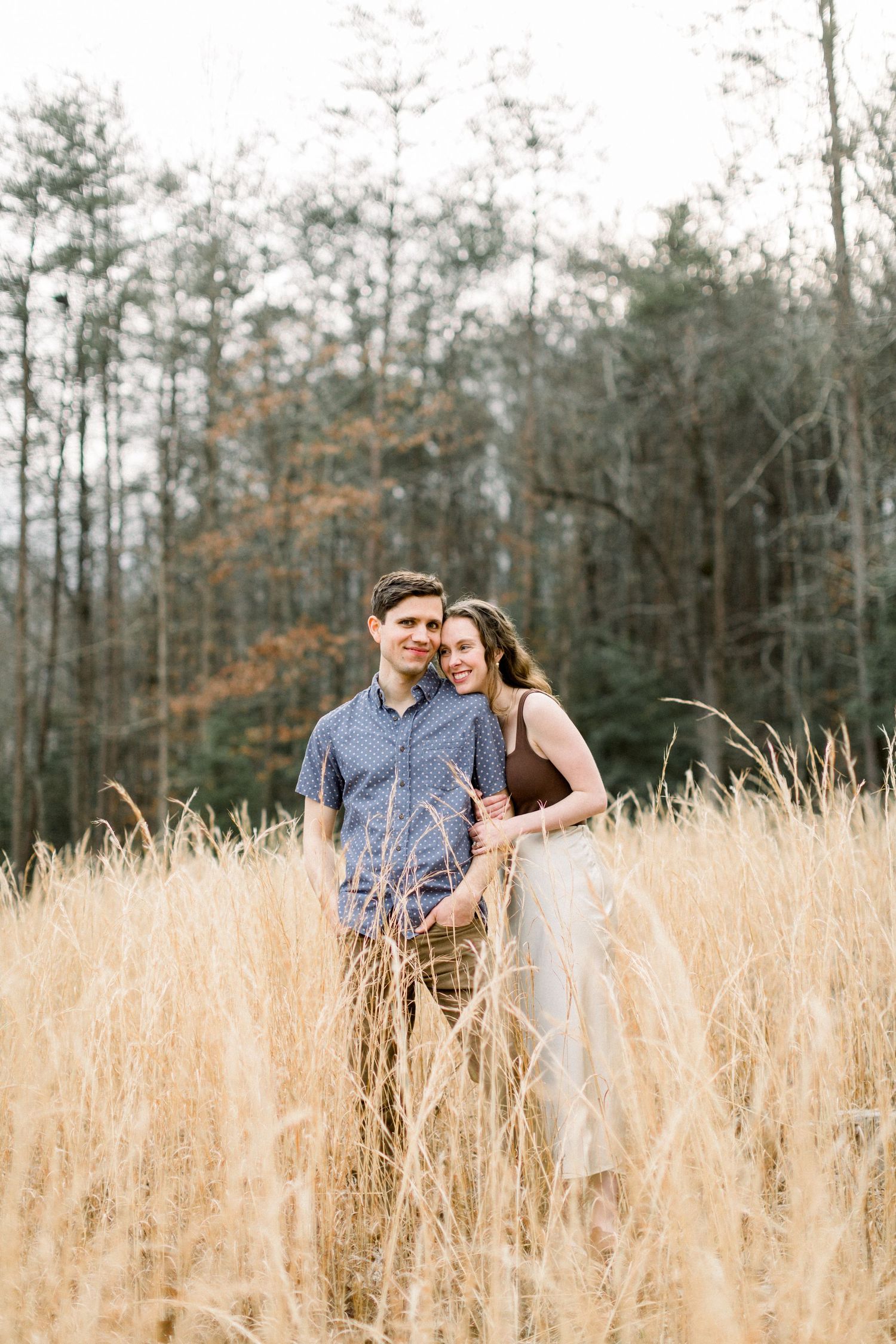 Happy couple embracing in a golden field of tall grass with autumn trees in the background.