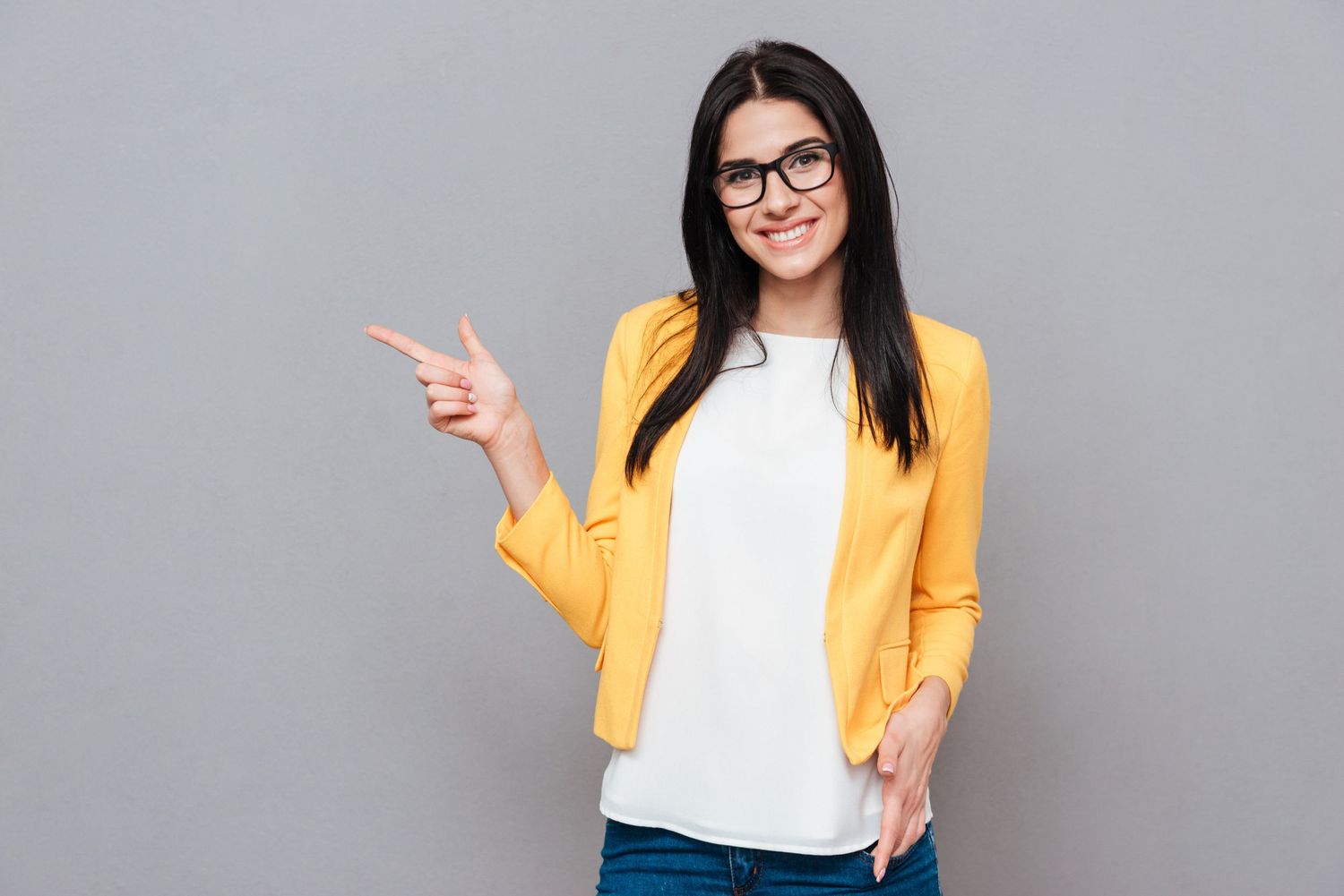 Person wearing glasses and yellow cardigan points to the side while smiling against grey background.