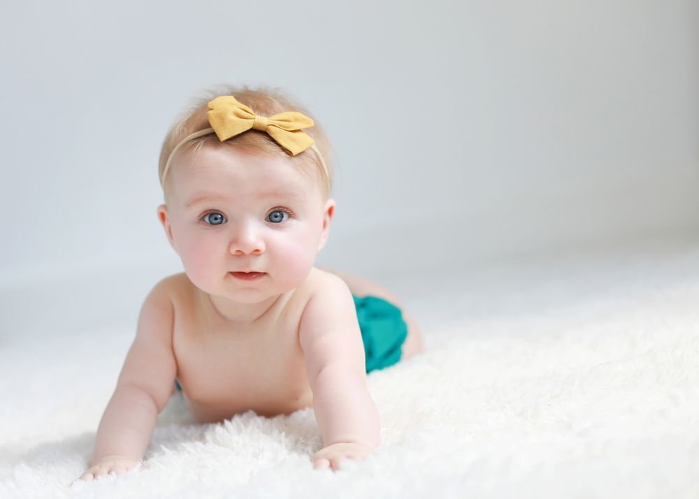 Baby lying on white fluffy rug wearing yellow bow headband.