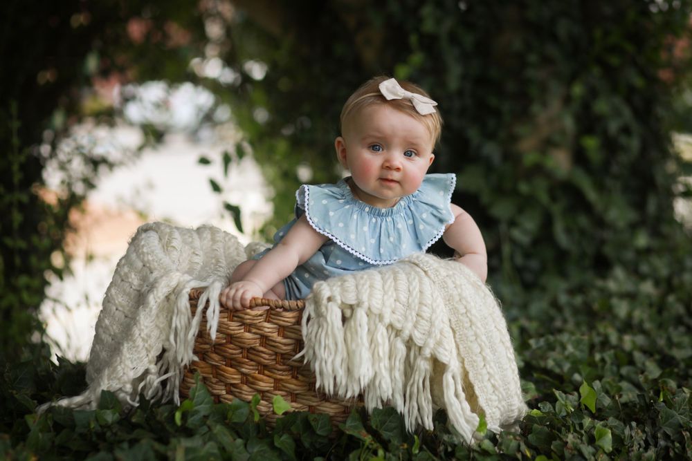 Sweet baby portrait in blue dress sitting in wicker basket with cream fringe blanket.