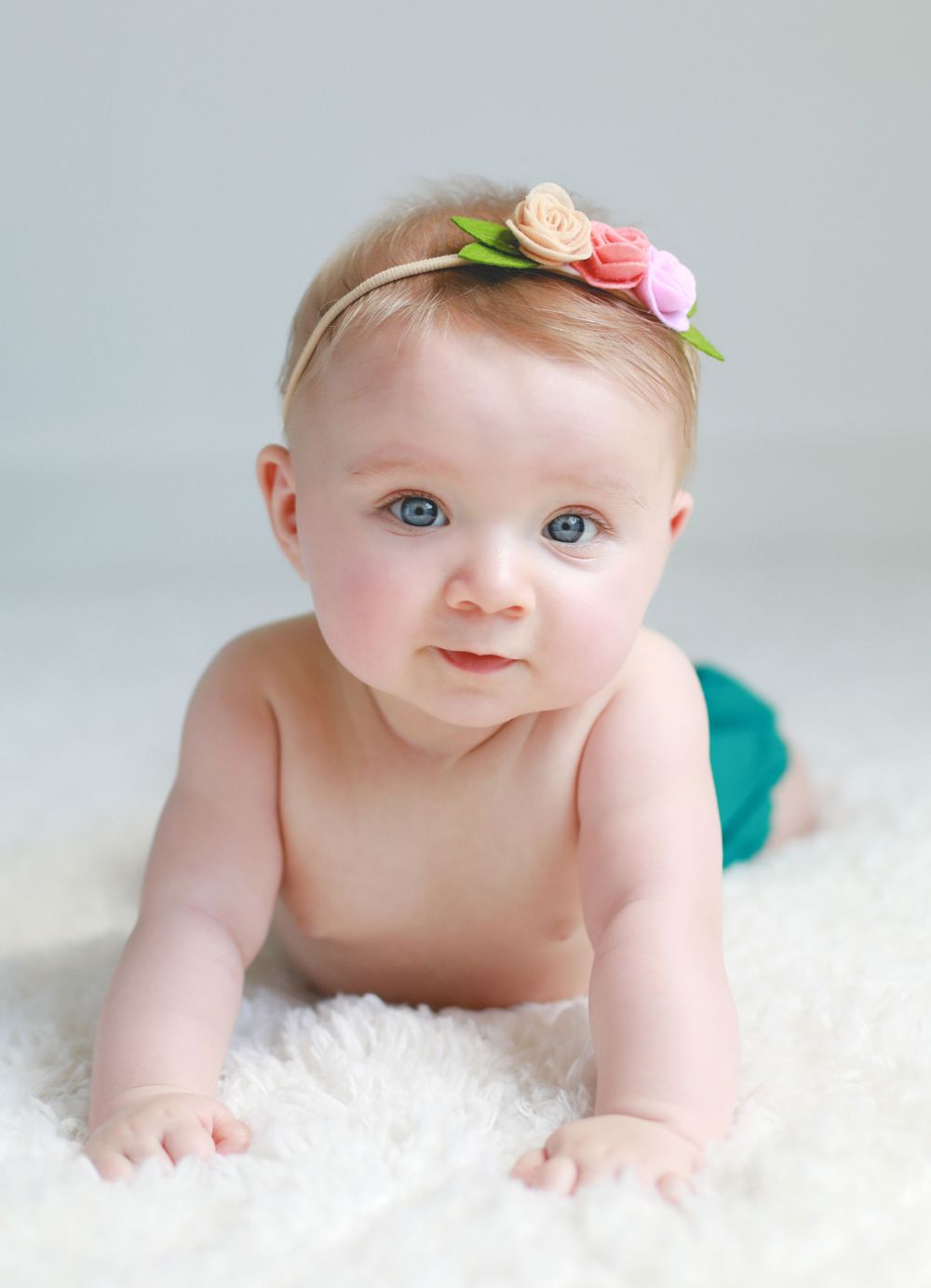 Baby on white fur rug wearing floral headband with soft pink and green details.