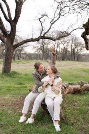 Two women smiling and holding hands while sitting on a fallen log in a bare winter woodland.