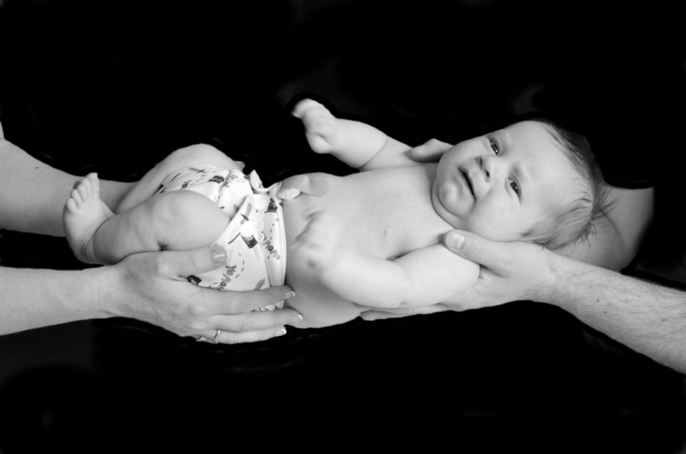 Black and white portrait of infant being held by adult hands against dark background.