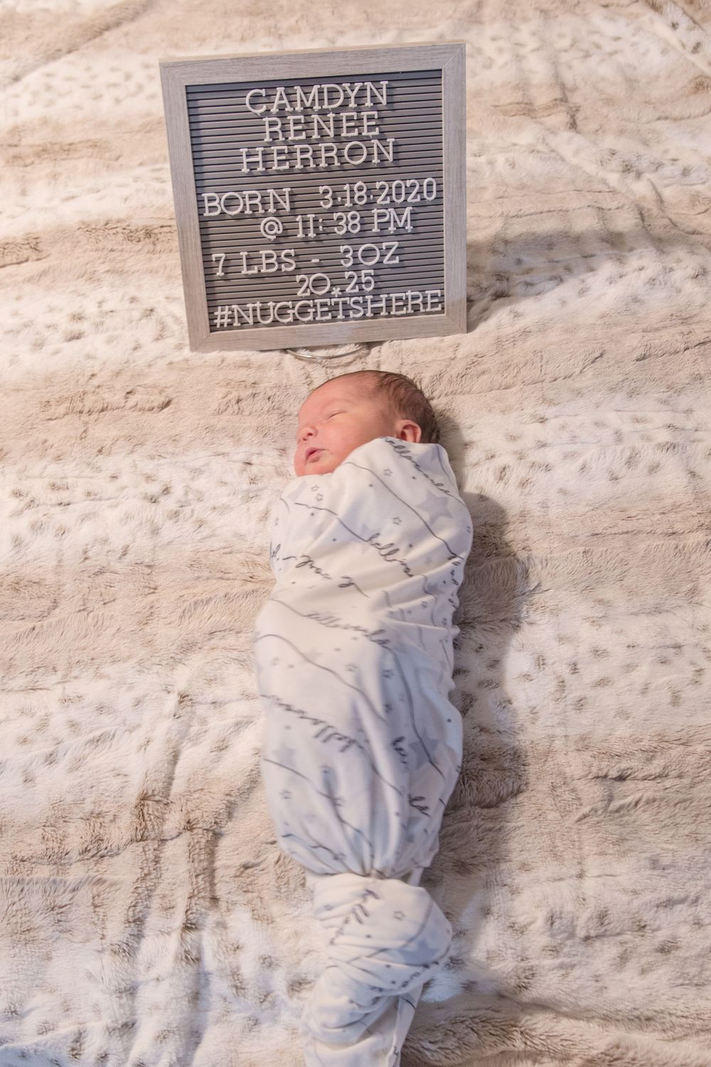 A newborn swaddled in white lies beneath a letterboard displaying birth details.