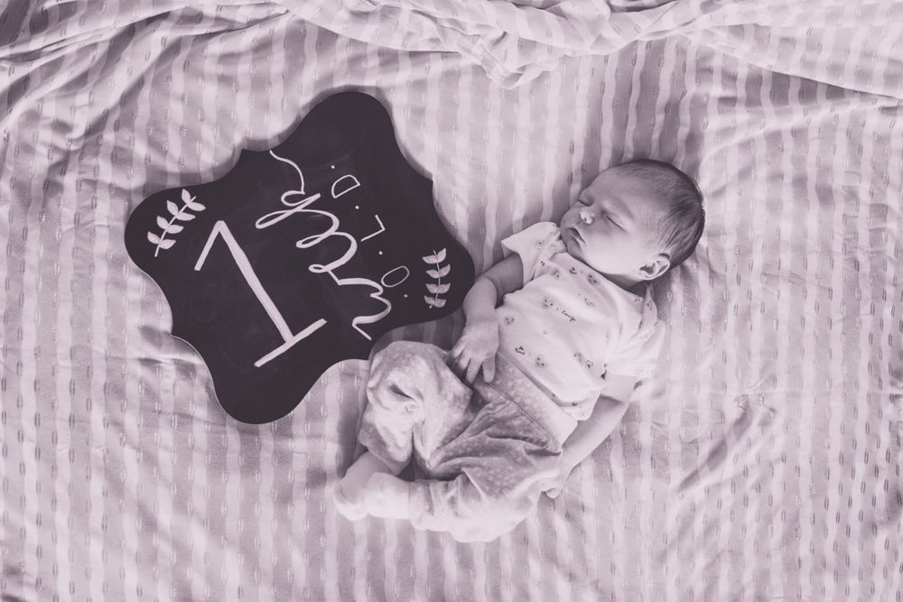 A swaddled newborn sleeps next to a black chalkboard milestone sign.