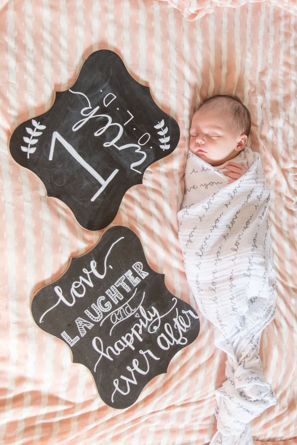 A sleeping newborn in white swaddle next to decorative chalkboard signs on striped bedding.