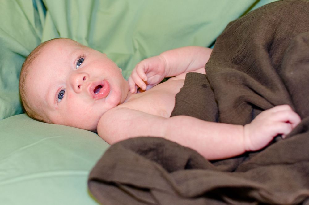 Baby lying on green bedding wrapped in brown blanket looking at camera with bright eyes.