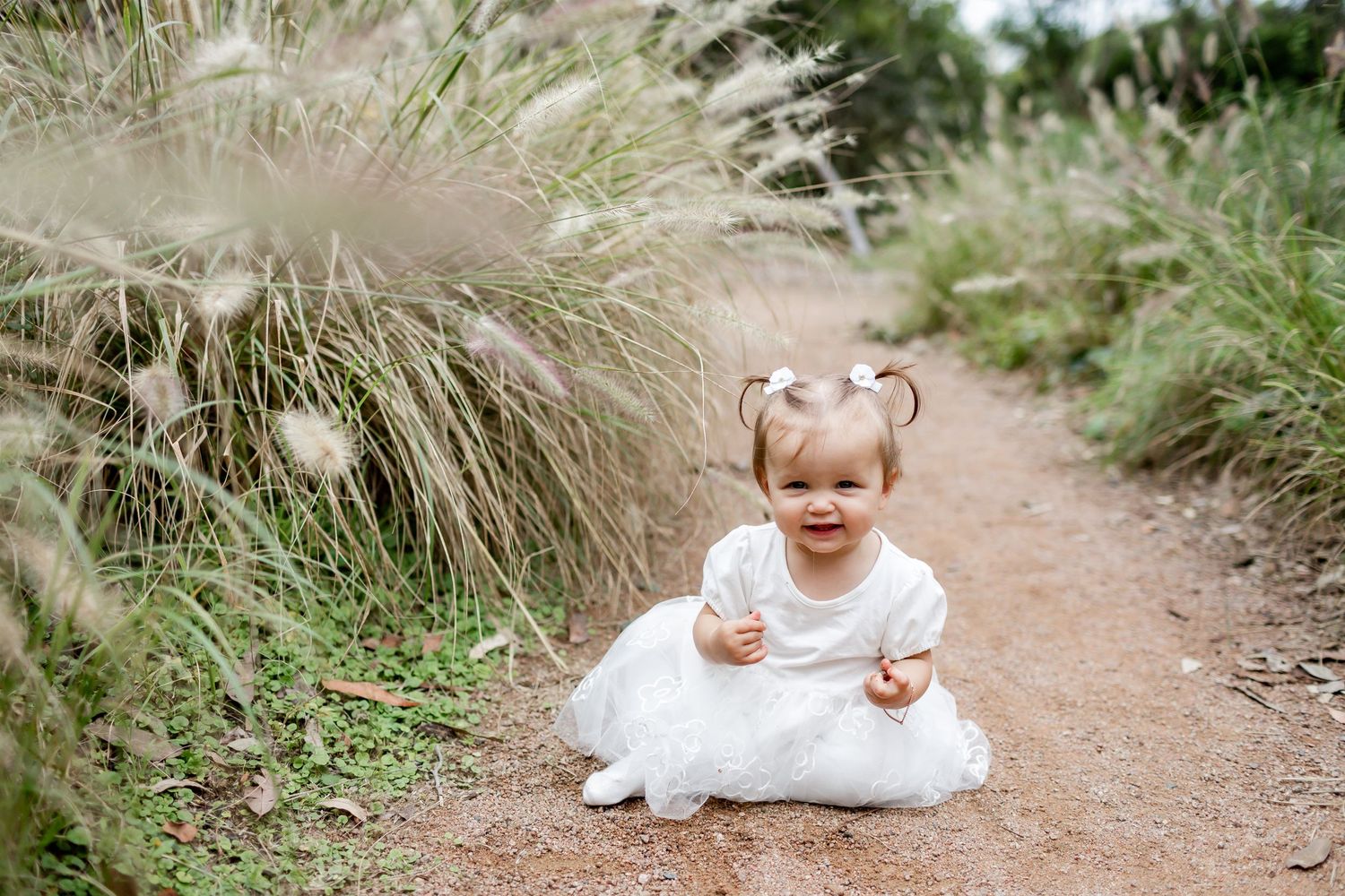 Joyful moment captured of child in white dress sitting on garden path surrounded by tall grasses.