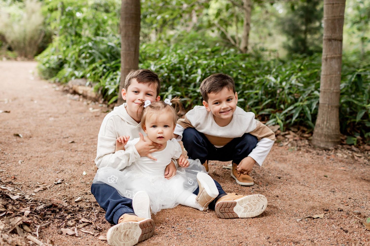 Three young children sitting together on a dirt path surrounded by lush garden foliage.