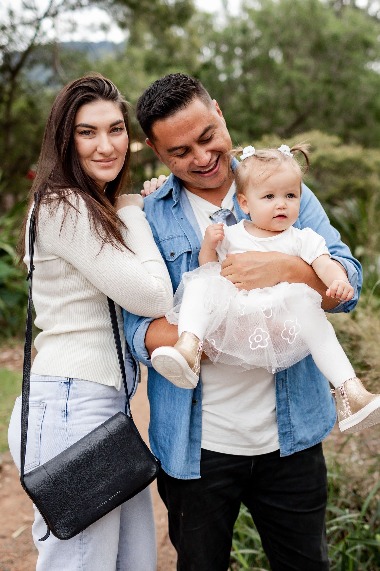 A couple in casual attire holds their baby girl wearing a white dress in an outdoor setting.