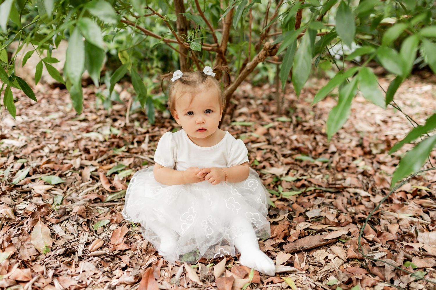Small child in a white dress sitting among garden mulch and leaves.