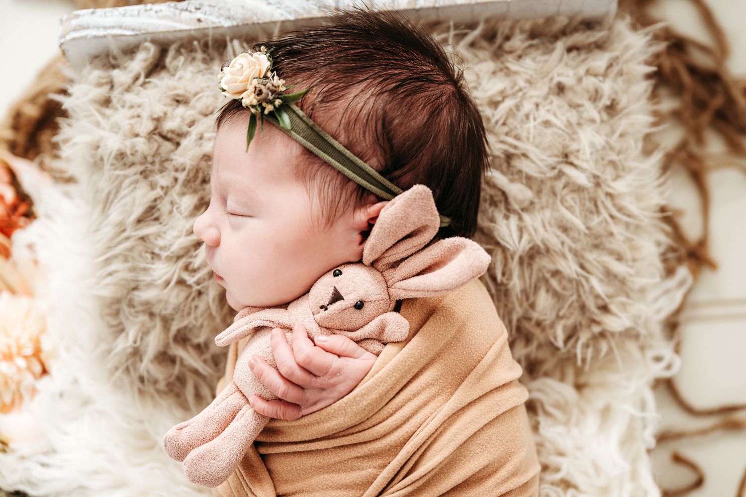 Sleeping newborn wrapped in beige fabric with floral headband on furry backdrop.