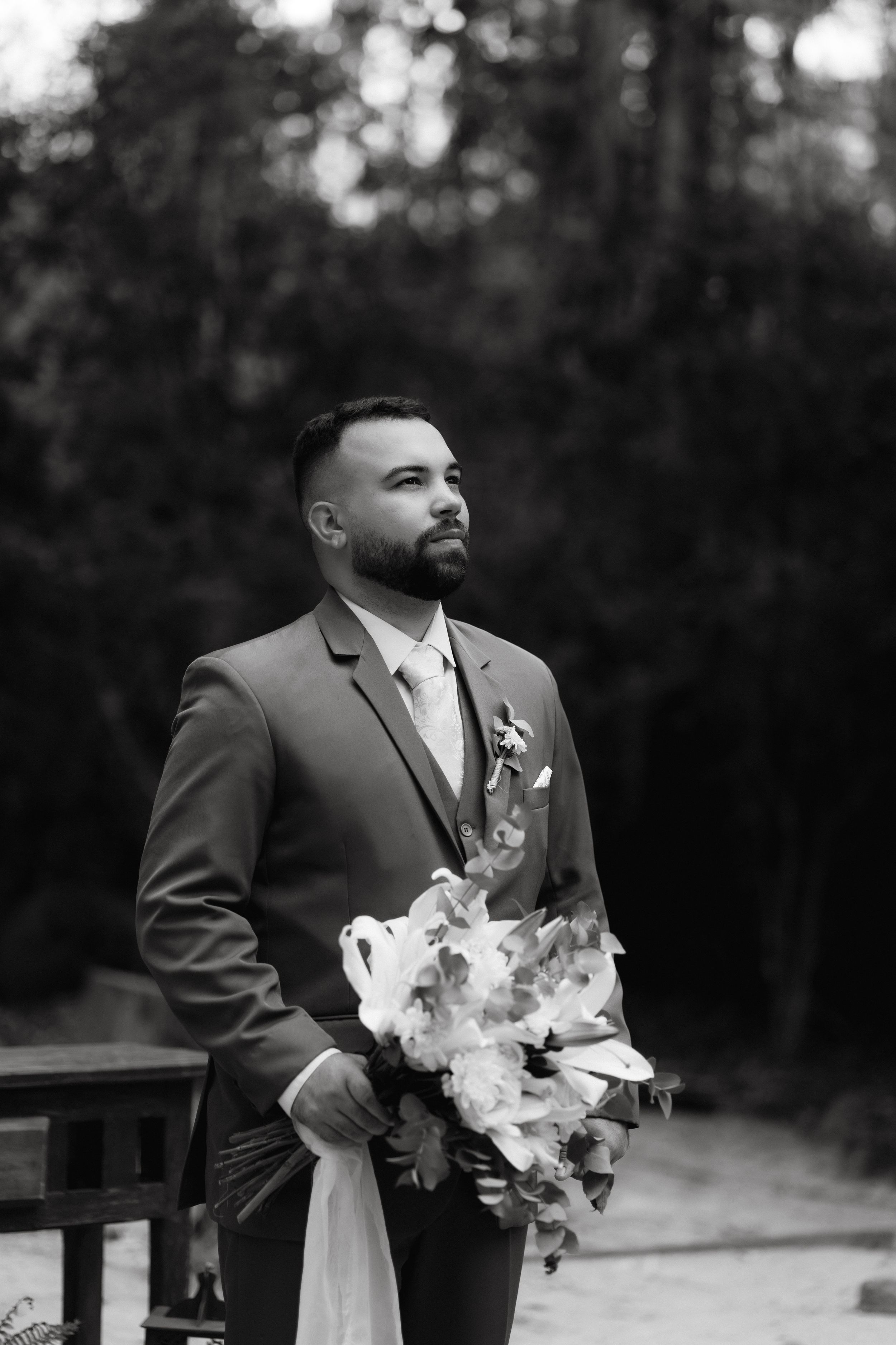 Black and white portrait of groom holding bouquet in forest setting.